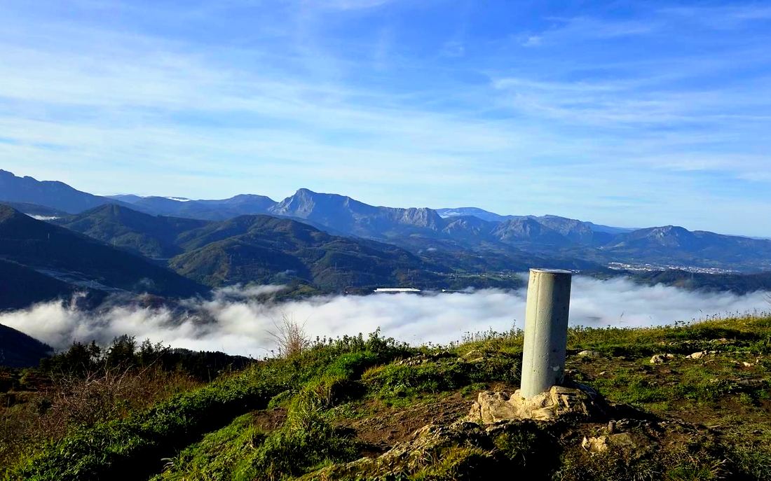 Vértice geodésico en la cumbre del Urko, con nubes bajas y las montañas del entorno al fondo
