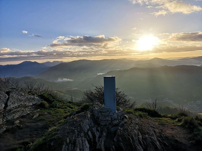 Urkoko gailurreko bertize geodesikoa ilunabarrean, harana eta herri bat atzealdean hodeien artean