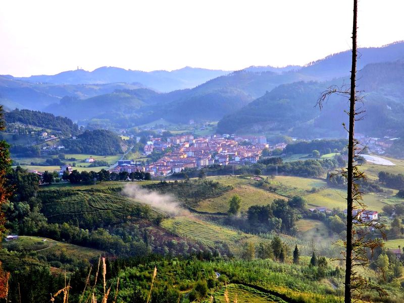 El pueblo en el fondo del valle al amanecer, visto desde lo alto, con los montes encadenados detr&aacute;s en tonos cada vez m&aacute;s claros