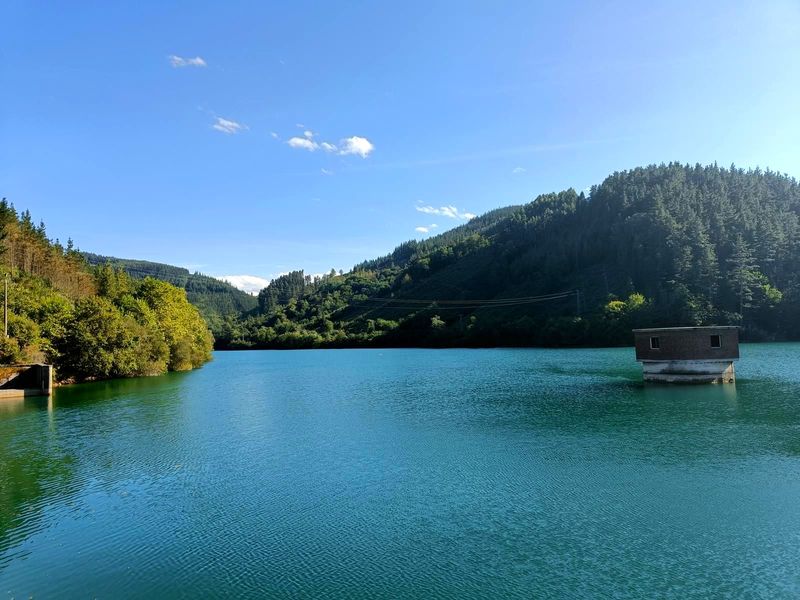 La presa de Aixola en un d&iacute;a soleado, con la torre de la toma de agua asomando sobre el embalse