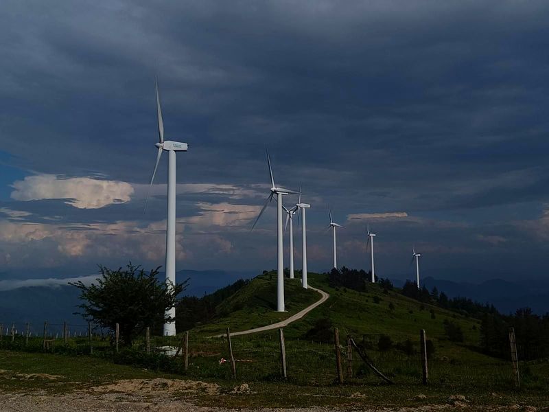Aerogeneradores en fila sobre una cresta de monte, con un cielo tormentoso al fondo